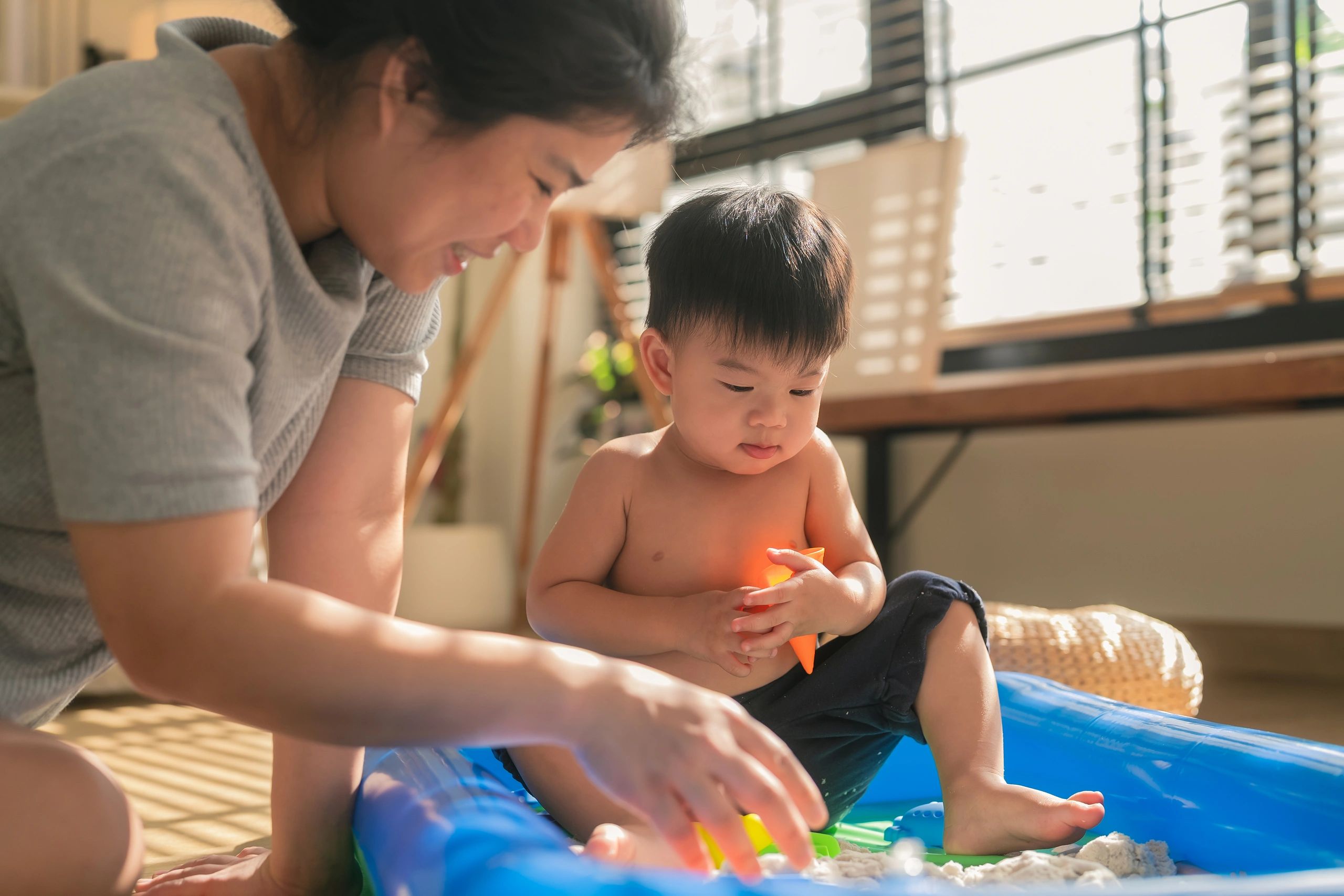 Asian mother and her toddler son enjoy a happy weekend together, playing with hydrophobic sand in their home. The little boy engages in sensory play in a home sandbox, helping develop fine motor skills an